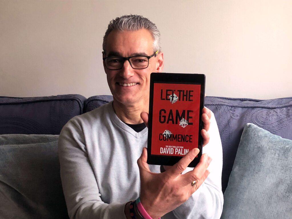 David Palin holds an e-reader displaying the cover of Let The Game Commence