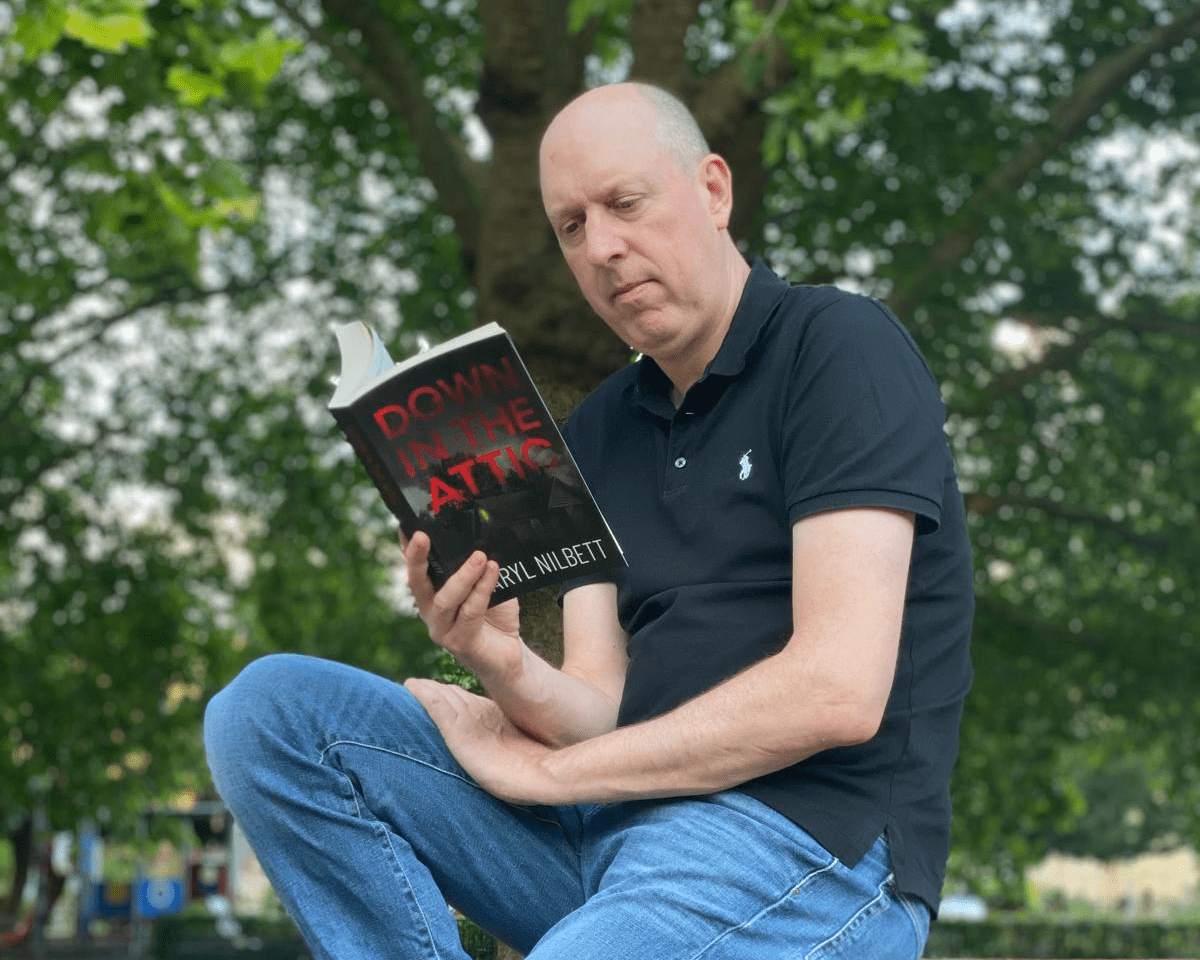 Photo of Down in the Attic author Daryl Nilbett reading his book outside on a bench.